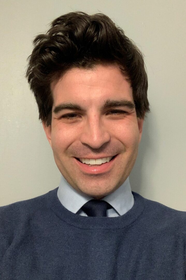 A man with a tie and sweater smiling for the camera.